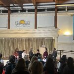 Mandy Moore and Cheryl Strayed sit on stage together at The Art of Living Retreat Center during a live storytelling session. The room is filled with attendees seated and listening intently. A projector displays a journaling exercise on screen, inviting participants to write a letter to their inner sage. Warm lighting, wooden beams, and golden curtains create a serene, sacred atmosphere. This moment captures Mandy sharing part of her healing journey alongside one of her literary heroes.