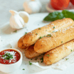 Freshly baked garlic breadsticks sprinkled with parsley and Parmesan, served with a side of marinara sauce on a white plate, with garlic cloves and tomatoes in the background.
