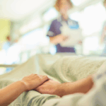 Close-up of a person lying in a hospital bed, holding hands with a loved one. In the blurred background, two nurses stand near the bed reviewing medical information. The image conveys tenderness, support, and the emotional weight of caregiving.