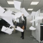 A woman in a professional outfit stands in a corporate office as sheets of paper dramatically fly through the air around her, symbolizing chaos, burnout, or a breaking point in a polished career setting.