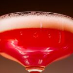 Close-up of a red sparkling cocktail in a coupe glass with a foamy head, symbolizing alcohol or celebratory drinking.