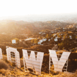 View from behind the iconic Hollywood sign at sunrise or sunset, overlooking the Los Angeles skyline and surrounding hills, with hazy golden light filling the scene.