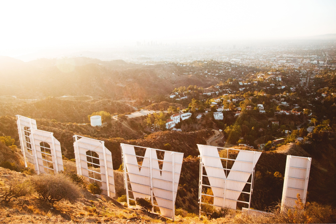 View from behind the iconic Hollywood sign at sunrise or sunset, overlooking the Los Angeles skyline and surrounding hills, with hazy golden light filling the scene.